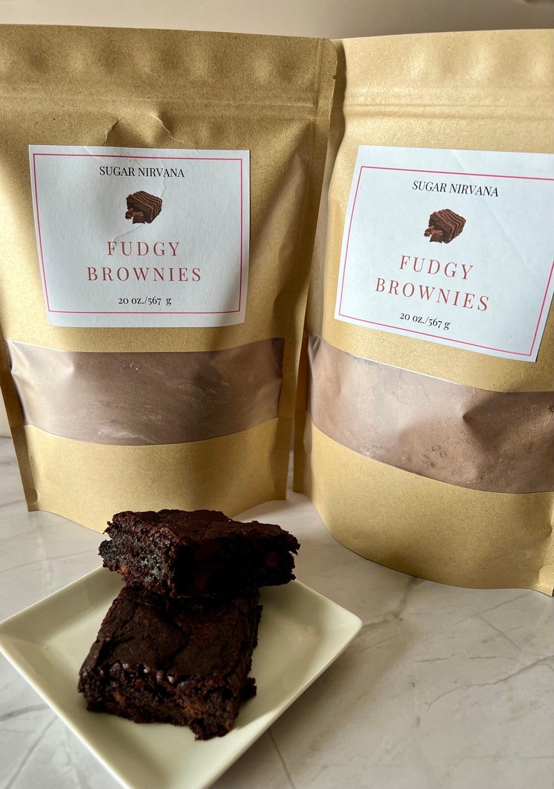 May include: Two brown paper bags with white labels that say "Sugar Nirvana" and "Fudgy Brownies" with a picture of a brownie on each label. The bags are filled with a brown powder. Two chocolate brownies are on a white plate.