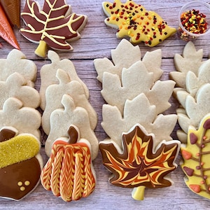 May include: An assortment of fall-themed cookies, including maple leaves, pumpkins, and acorns, decorated with various colors and sprinkles. The cookies are arranged on a wooden surface, suggesting a seasonal treat.