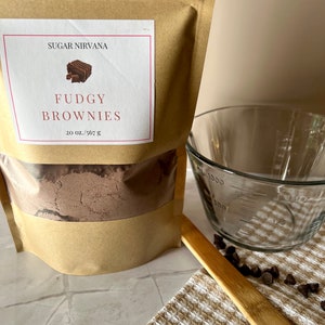 May include: A brown paper bag with a label that reads "Sugar Nirvana Fudgy Brownies 20 oz./567 g". The bag contains a brown powder. A glass measuring cup, a wooden spoon, and chocolate chips are also in the image.