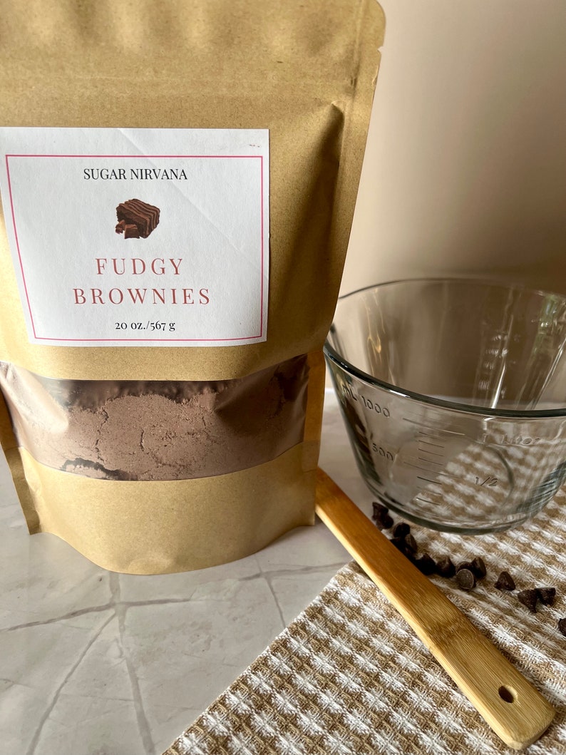 May include: A brown paper bag with a label that reads "Sugar Nirvana Fudgy Brownies 20 oz./567 g". The bag contains a brown powder. A glass measuring cup, a wooden spoon, and chocolate chips are also in the image.