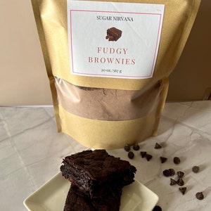 May include: A brown paper bag with a white label that reads "Sugar Nirvana Fudgy Brownies 20 oz./567 g". Two chocolate brownies are stacked on a white square plate. Chocolate chips are scattered around the plate.