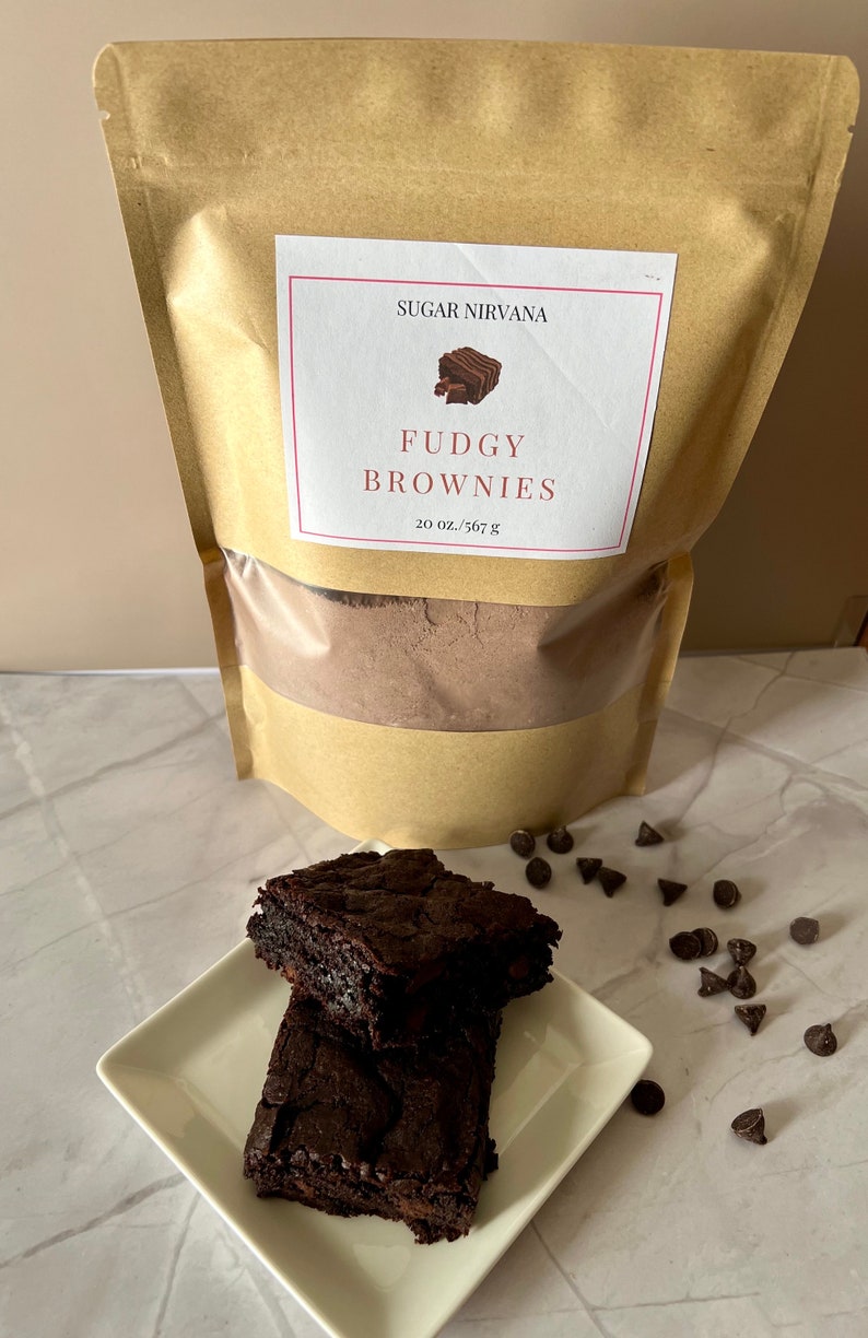 May include: A brown paper bag with a white label that reads "Sugar Nirvana Fudgy Brownies 20 oz./567 g". Two chocolate brownies are stacked on a white square plate. Chocolate chips are scattered around the plate.
