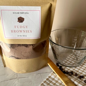 May include: A brown paper bag with a white label that says "Sugar Nirvana" and "Fudgy Brownies 20 oz./567 g". The bag contains a brown powder. There is a glass measuring cup, a wooden spoon, and chocolate chips on a brown and white checkered towel.