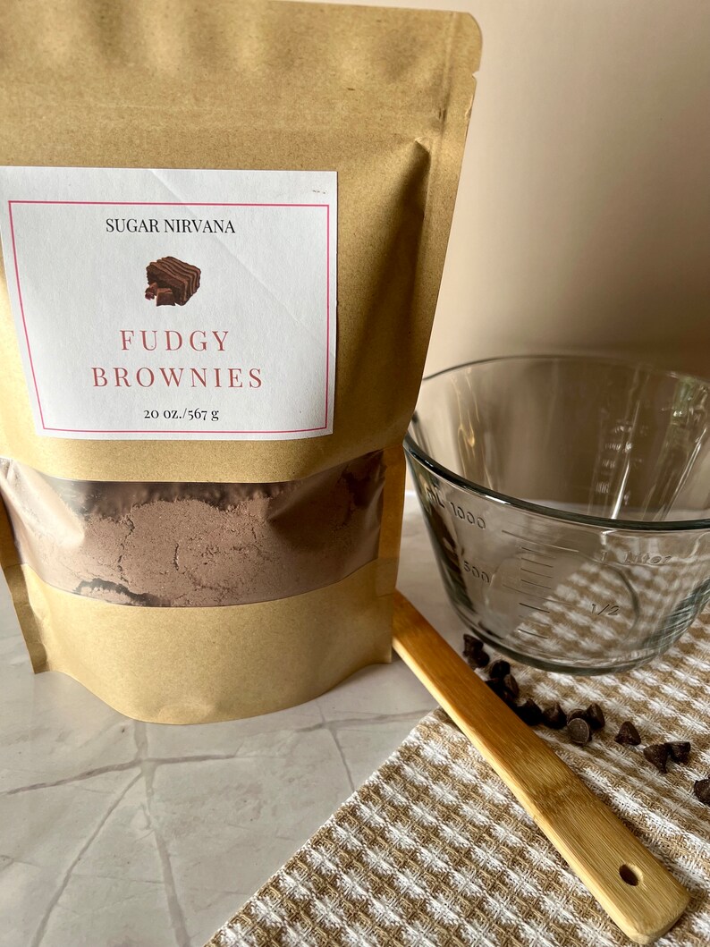 May include: A brown paper bag with a white label that says "Sugar Nirvana" and "Fudgy Brownies 20 oz./567 g". The bag contains a brown powder. There is a glass measuring cup, a wooden spoon, and chocolate chips on a brown and white checkered towel.