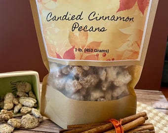 May include: A brown paper bag with a label that reads "Sugar Nirvana's Candied Cinnamon Pecans 1 lb. (453 Grams)". The bag is filled with candied pecans. A small bowl of candied pecans is in the foreground, along with a bundle of cinnamon sticks.