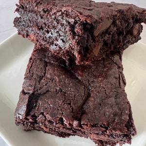 May include: Two squares of fudgy chocolate brownies on a white plate. The brownies are dark brown and have a slightly cracked surface.