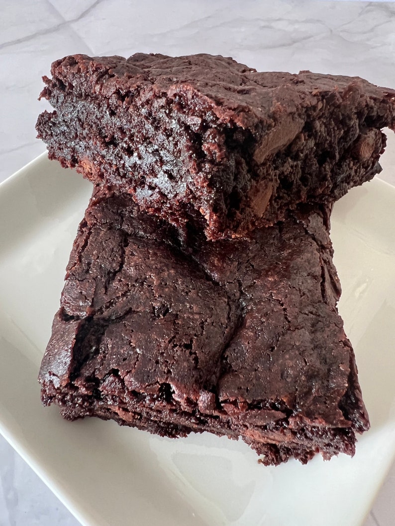 May include: Two squares of fudgy chocolate brownies on a white plate. The brownies are dark brown and have a slightly cracked surface.