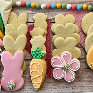 May include: A set of Easter-themed sugar cookies with various designs, including bunnies, carrots, flowers, and eggs. The cookies are decorated with pink, orange, and green icing and sprinkles. The set also includes bags of sprinkles and icing.