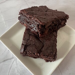 May include: Two chocolate brownies on a white square plate. The brownies are dark brown and have a slightly crispy texture.
