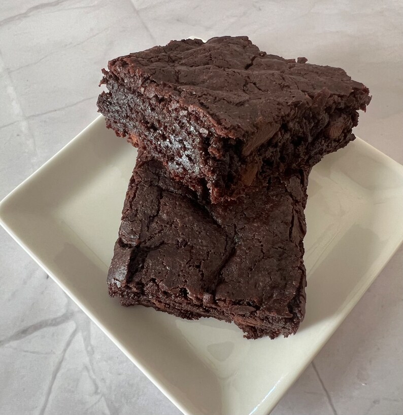 May include: Two chocolate brownies on a white square plate. The brownies are dark brown and have a slightly crispy texture.