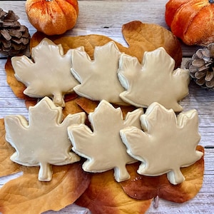May include: Six maple leaf shaped sugar cookies with brown icing on a wooden surface with fall-colored leaves and two orange pumpkins.