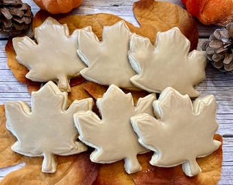 May include: Six maple leaf shaped sugar cookies with brown icing on a wooden surface with fall-colored leaves and two orange pumpkins.