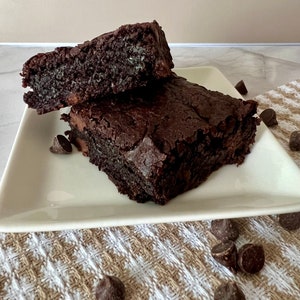 May include: Two chocolate brownies on a white plate. The brownies are dark brown and have a slightly crispy texture. There are chocolate chips scattered around the plate.