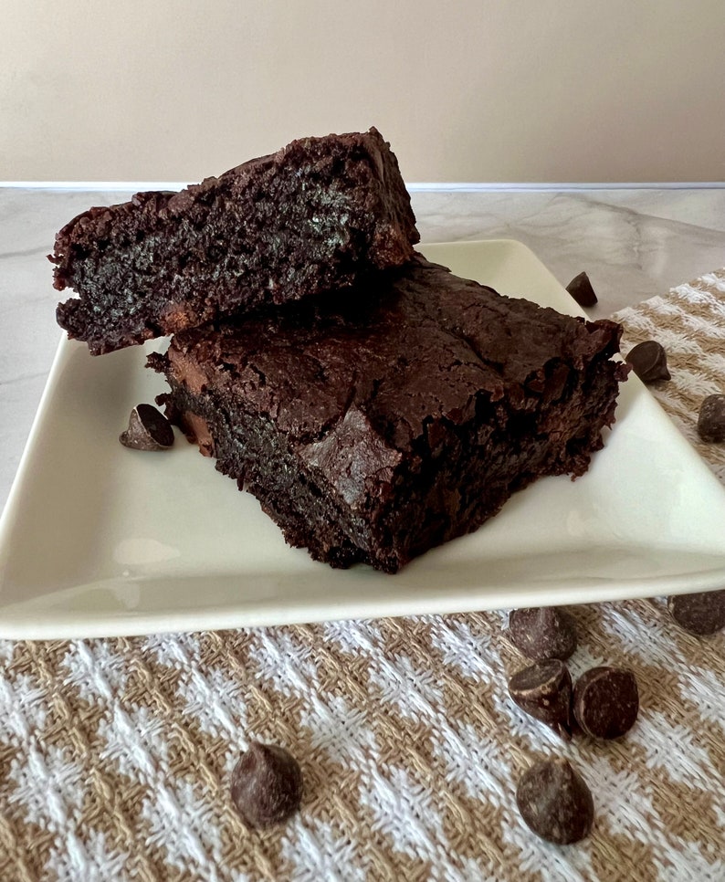 May include: Two chocolate brownies on a white plate. The brownies are dark brown and have a slightly crispy texture. There are chocolate chips scattered around the plate.
