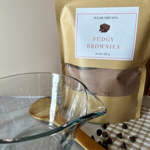 May include: A brown paper bag with a white label that says "Sugar Nirvana" and "Fudgy Brownies" with a picture of a brownie. The bag contains a brown powder. There is a glass measuring cup, a wooden spoon, and chocolate chips on a white and brown checkered towel.