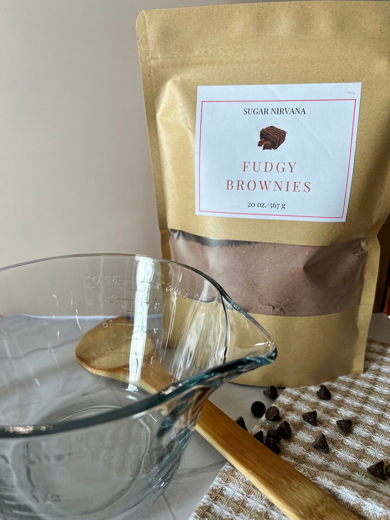 May include: A brown paper bag with a white label that says "Sugar Nirvana" and "Fudgy Brownies" with a picture of a brownie. The bag contains a brown powder. There is a glass measuring cup, a wooden spoon, and chocolate chips on a white and brown checkered towel.