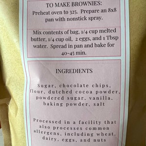 May include: A pink and white label with instructions on how to make brownies. The ingredients include sugar, chocolate chips, flour, cocoa powder, powdered sugar, vanilla, baking powder, and salt. The label also states that the product is processed in a facility that also processes common allergens, including wheat, dairy, eggs, and nuts.