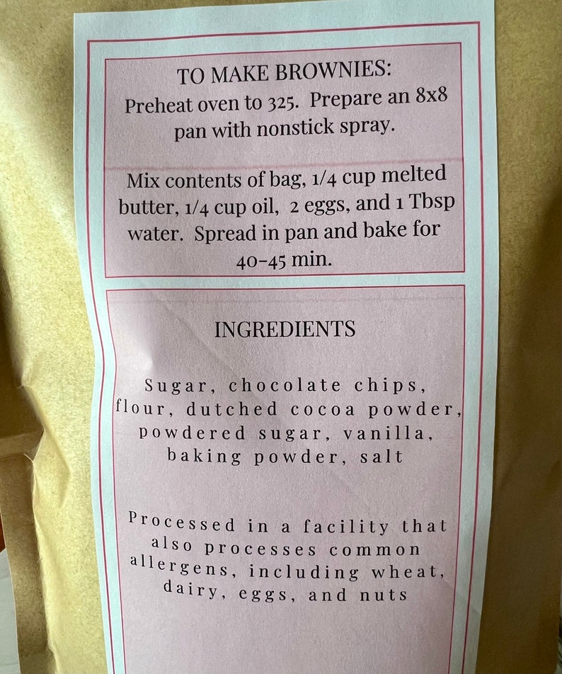 May include: A pink and white label with instructions on how to make brownies. The ingredients include sugar, chocolate chips, flour, cocoa powder, powdered sugar, vanilla, baking powder, and salt. The label also states that the product is processed in a facility that also processes common allergens, including wheat, dairy, eggs, and nuts.