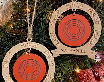 May include: Two wooden Christmas ornaments with a clay shooting target design. The ornaments are brown with the text "Break Clay" repeated around the edge. The top ornament has the name "Nathaniel" and the year "2023" on the bottom. The bottom ornament has the year "2023" on the bottom.