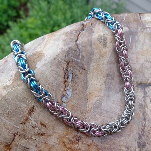 May include: A blue and pink chainmail necklace made of metal rings. The necklace is lying on a textured rock surface.