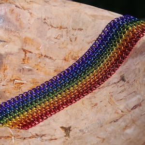 Pode incluir: Um bracelete de malha de corrente arco-íris feito de pequenos anéis de metal. O bracelete é feito nas cores do arco-íris, com cada cor representada por uma fila de anéis.