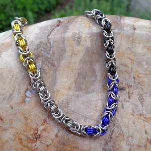 May include: A silver chainmail necklace with yellow, black, and blue accents.