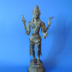 May include: Bronze statue of a deity with multiple arms, adorned with intricate details. The figure stands on a decorative base against a blue backdrop. The statue's attire includes a crown and draped garments, showcasing a traditional aesthetic.