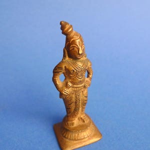May include: A small, bronze-colored statue of a figure with arms akimbo, standing on a square base. The figure has detailed facial features and wears a headdress. The statue is likely a religious or decorative item.