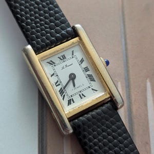 Peut inclure: Une montre-bracelet rectangulaire dor&eacute;e avec un cadran blanc et des chiffres romains noirs. La montre a un bracelet en cuir noir. Le cadran de la montre indique "Le Grand" et "17 Jewels".