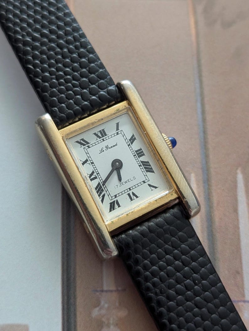 Peut inclure: Une montre-bracelet rectangulaire dor&eacute;e avec un cadran blanc et des chiffres romains noirs. La montre a un bracelet en cuir noir. Le cadran de la montre indique "Le Grand" et "17 Jewels".