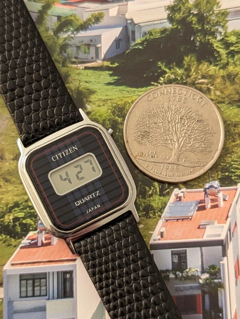 May include: A vintage Citizen digital watch with a black textured band and a silver-tone case. The watch face displays the time as 4:27. A Connecticut quarter coin is next to the watch.