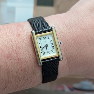 Peut inclure: Une montre-bracelet rectangulaire dor&eacute;e avec un cadran blanc et des chiffres romains noirs. La montre a un bracelet en cuir noir textur&eacute;. Le cadran de la montre indique "17 Jewels" et "Le Manuel". Une petite gemme bleue est sur le c&ocirc;t&eacute; de la montre.