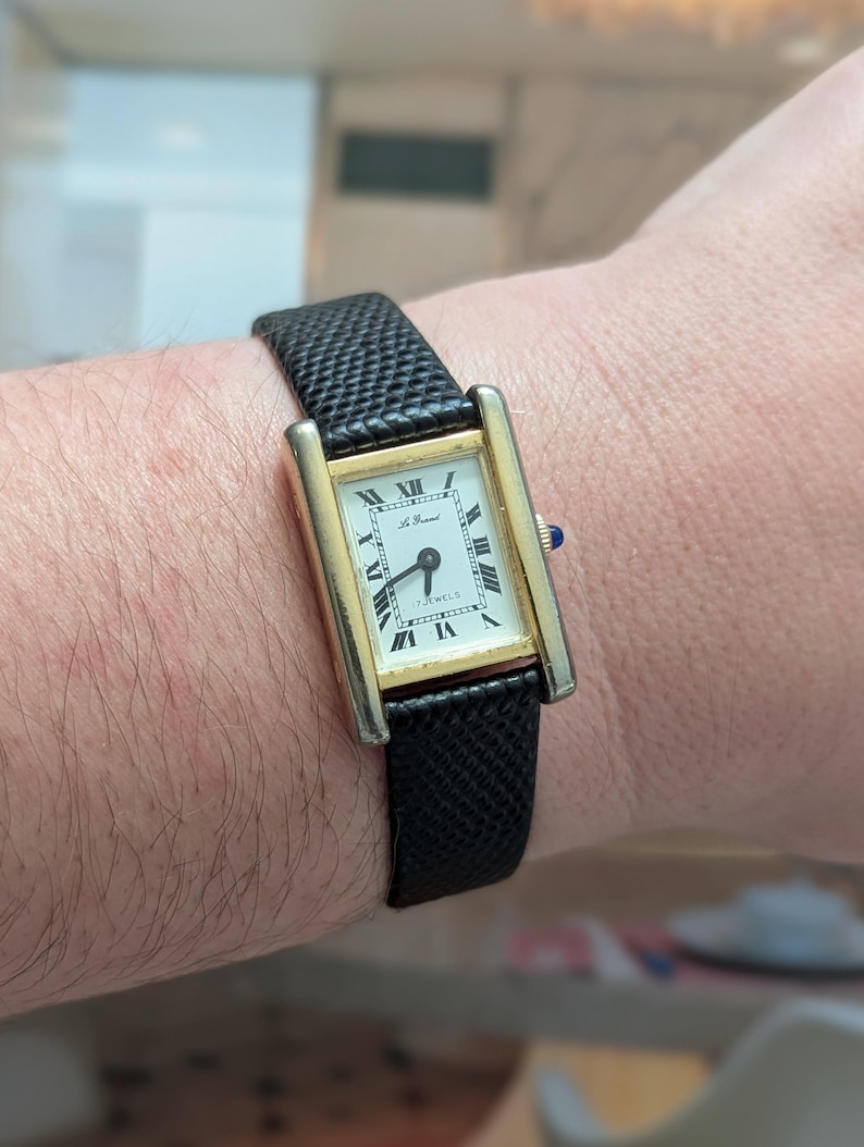 Peut inclure: Une montre-bracelet rectangulaire dor&eacute;e avec un cadran blanc et des chiffres romains noirs. La montre a un bracelet en cuir noir textur&eacute;. Le cadran de la montre indique "17 Jewels" et "Le Manuel". Une petite gemme bleue est sur le c&ocirc;t&eacute; de la montre.
