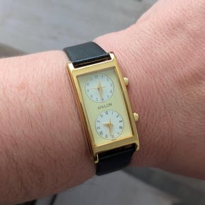 Puede incluir: Reloj rectangular dorado con dos esferas color crema y una correa de cuero negro. El reloj muestra la marca "AVALON" y n&uacute;meros romanos. El reloj se lleva en la mu&ntilde;eca.