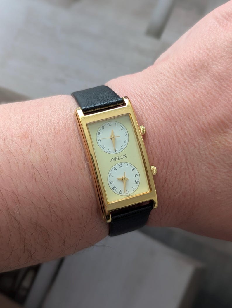 Puede incluir: Reloj rectangular dorado con dos esferas color crema y una correa de cuero negro. El reloj muestra la marca "AVALON" y n&uacute;meros romanos. El reloj se lleva en la mu&ntilde;eca.