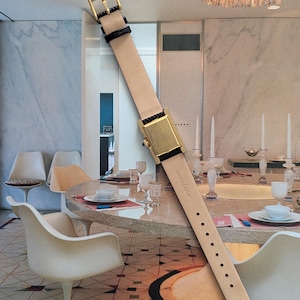 Peut inclure: Une montre-bracelet rectangulaire dor&eacute;e avec un bracelet en cuir beige et des d&eacute;tails noirs. La montre a une boucle dor&eacute;e et se d&eacute;tache sur un arri&egrave;re-plan int&eacute;rieur flou. Le bracelet est marqu&eacute; "Genuine Leather" et porte le num&eacute;ro "15".