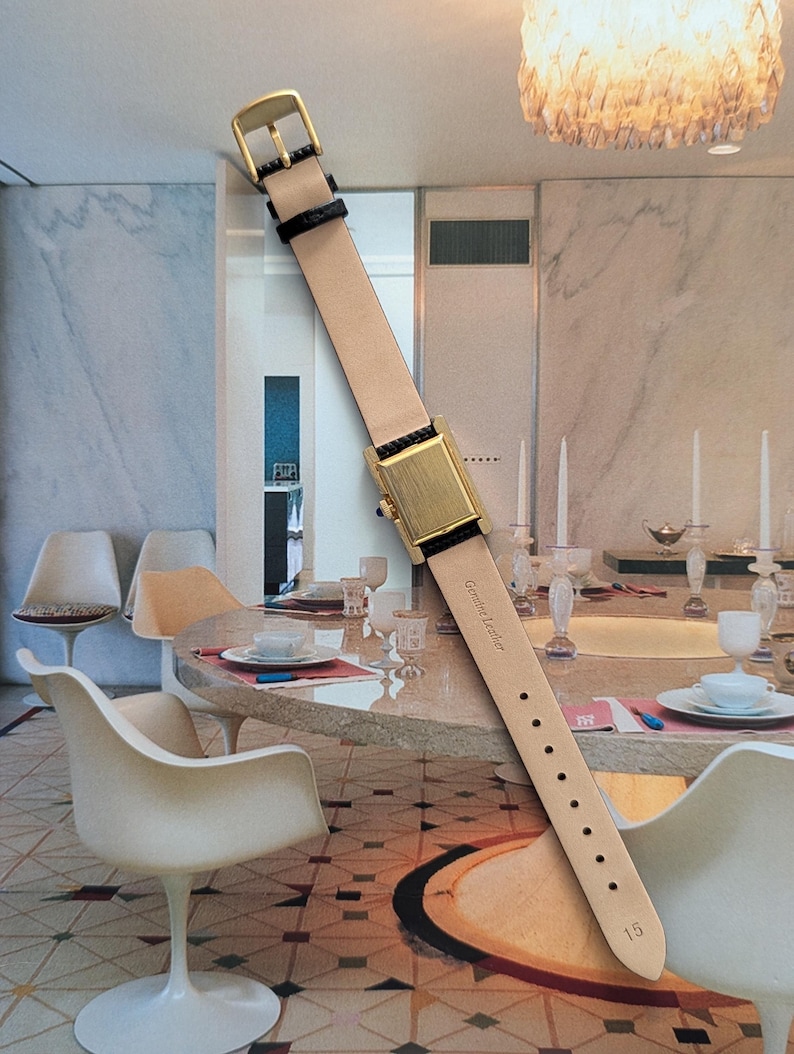 Peut inclure: Une montre-bracelet rectangulaire dor&eacute;e avec un bracelet en cuir beige et des d&eacute;tails noirs. La montre a une boucle dor&eacute;e et se d&eacute;tache sur un arri&egrave;re-plan int&eacute;rieur flou. Le bracelet est marqu&eacute; "Genuine Leather" et porte le num&eacute;ro "15".