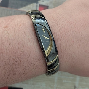 May include: A Seiko Rivoli wristwatch featuring a black teardrop-shaped dial with gold-tone hands and markers. The watch has a wide, integrated bracelet band made of alternating black and gold-tone curved metal segments that wrap around the wrist.