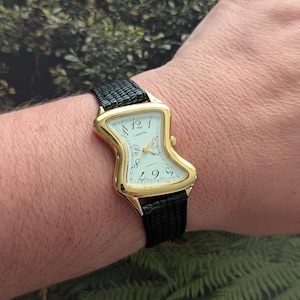 Puede incluir: Un reloj de pulsera dorado con una caja curvada en forma de reloj de arena y una esfera blanca con números negros y el texto Capezio. El reloj tiene manecillas doradas y una correa de cuero negro texturizado con un patrón de piel de lagarto.