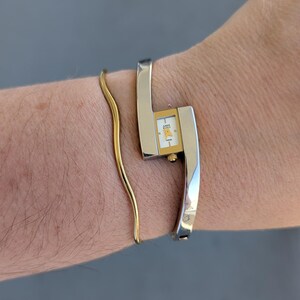 Puede incluir: Un reloj tipo brazalete plateado con esfera rectangular blanca, manecillas doradas y el logotipo de Anne Klein, junto a una pulsera fina de alambre dorado ondulado. El reloj tiene un acabado metálico pulido y una pequeña corona dorada lateral.