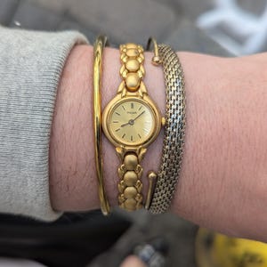 May include: A gold-tone Pulsar wristwatch with a round face and a gold-tone metal band. The watch is worn on a wrist with three gold-tone bracelets.