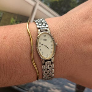 Può includere: Un orologio Seiko Quartz color argento con quadrante ovale color crema, numeri arabi neri e cinturino in metallo a maglie multiple, accompagnato da un bracciale rigido in metallo dorato sottile e ondulato.