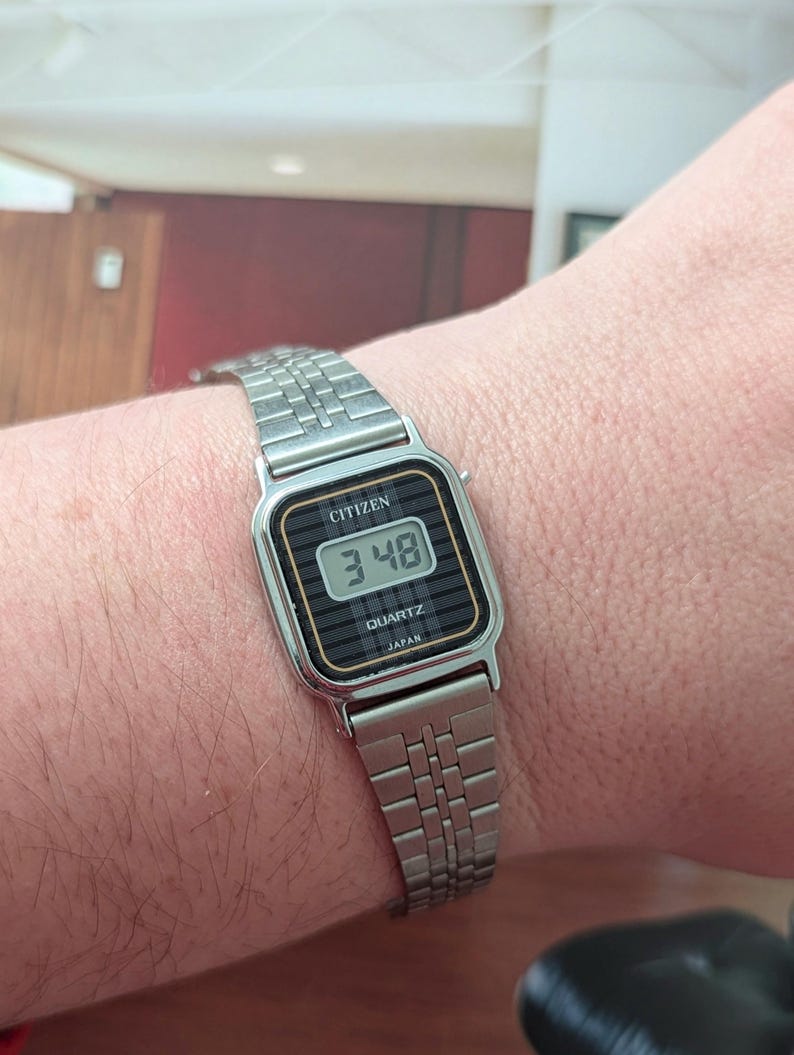 May include: A silver Citizen Quartz digital watch displaying the time 3:48. The watch has a black face with white numerals and a silver metal band. The watch is rectangular with rounded corners and the word "Japan" is printed on the watch face.