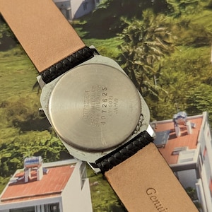 May include: A vintage Citizen watch with a silver-toned case and a black leather strap. The watch back is visible, displaying the brand name and model details. The strap is tan on the inside and embossed with "Genuine Leather".