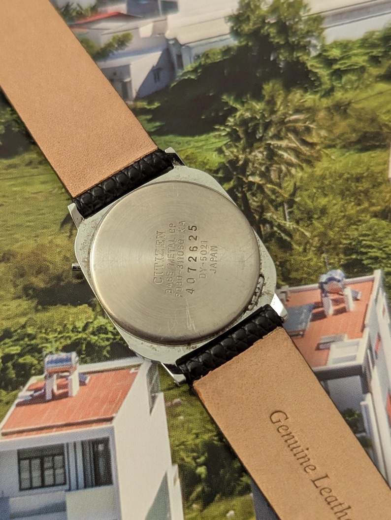 May include: A vintage Citizen watch with a silver-toned case and a black leather strap. The watch back is visible, displaying the brand name and model details. The strap is tan on the inside and embossed with "Genuine Leather".