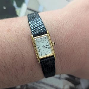 May include: A gold-coloured rectangular Pulsar Quartz watch with a black textured leather strap. The watch face is white with black Roman numerals and hands. The watch is worn on a wrist.