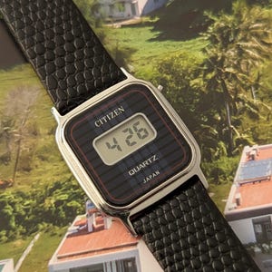 May include: A Citizen Quartz digital watch with a black face and a black textured leather band. The watch displays the time as 4:26. The watch face has a silver-colored metal frame. The watch is labeled "JAPAN".