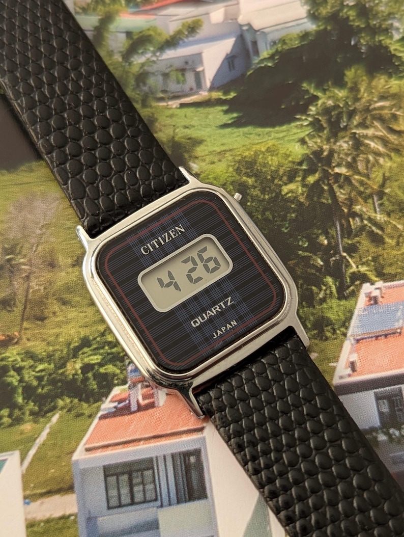 May include: A Citizen Quartz digital watch with a black face and a black textured leather band. The watch displays the time as 4:26. The watch face has a silver-colored metal frame. The watch is labeled "JAPAN".