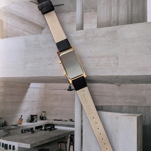 Puede incluir: Un reloj rectangular dorado con una correa de cuero negro y una parte inferior de color crema. El reloj tiene una hebilla dorada y una esfera plateada. La correa est&aacute; etiquetada como "Genuine Leather".