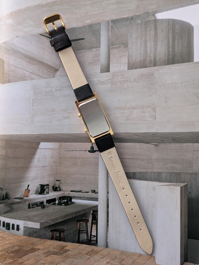 Puede incluir: Un reloj rectangular dorado con una correa de cuero negro y una parte inferior de color crema. El reloj tiene una hebilla dorada y una esfera plateada. La correa est&aacute; etiquetada como "Genuine Leather".